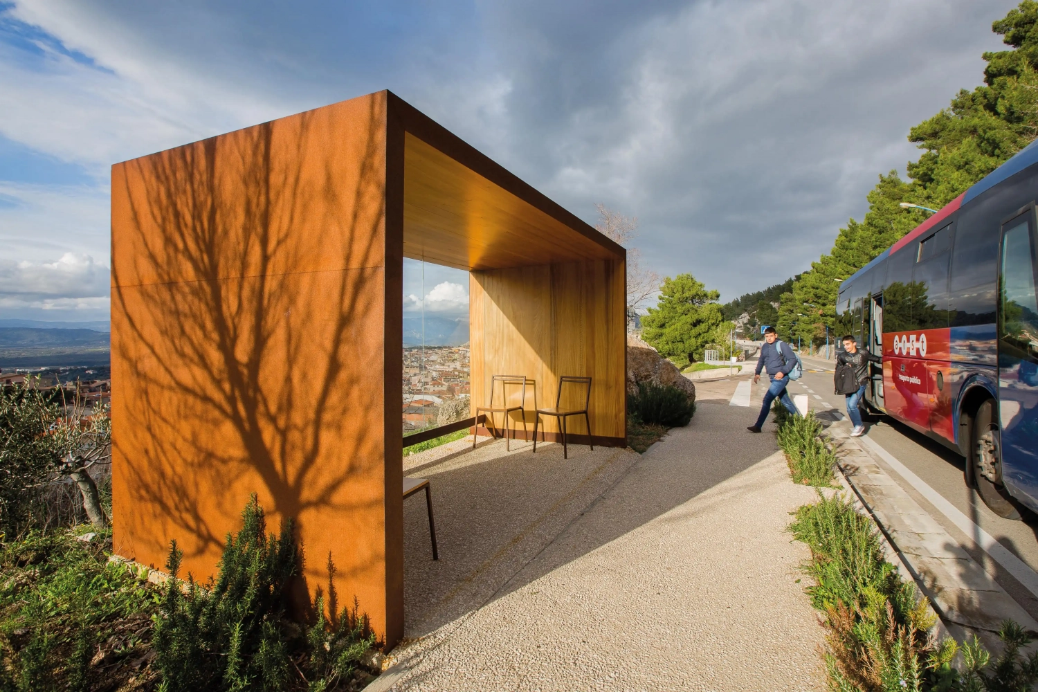 Moderne Überdachung aus rostfreiem Stahl und Holz, umgeben von Landschaft, mit zwei Stühlen innen und einer Person, die vorbeigeht.
