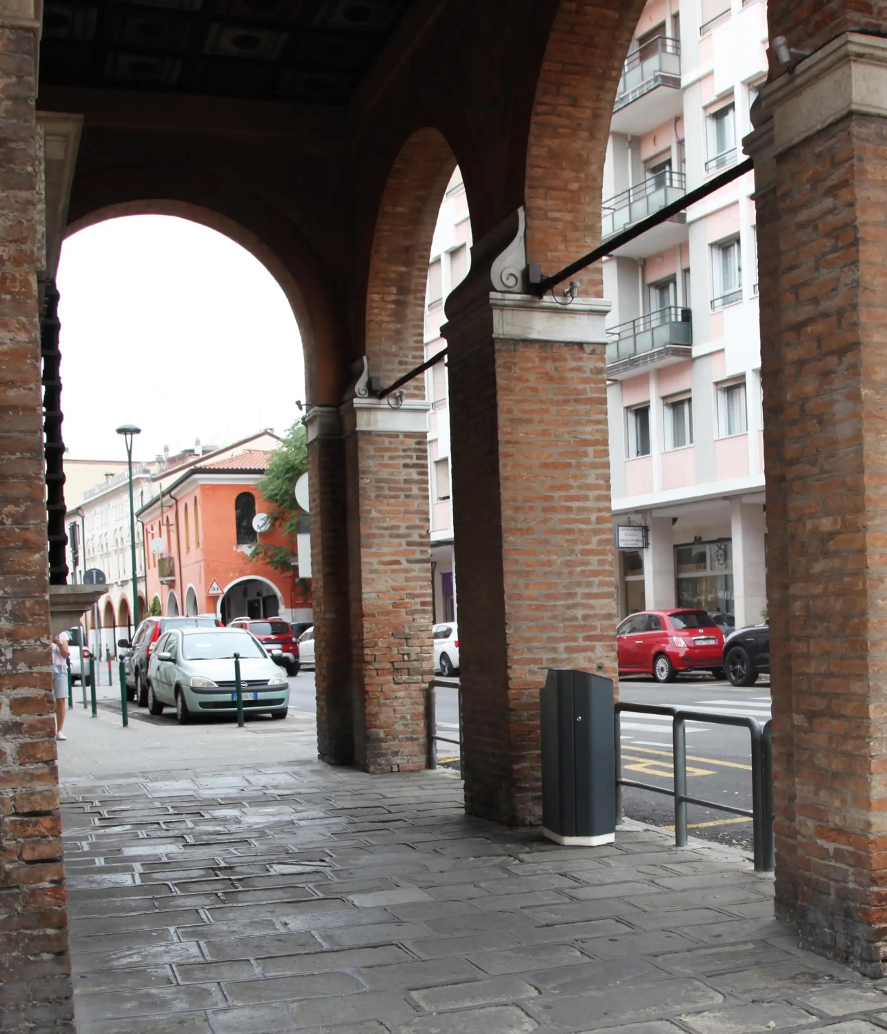 Abfallbehälter steht unter einem Bogen in einer Stadtstraße, umgeben von Ziegelarchitektur und geparkten Autos.