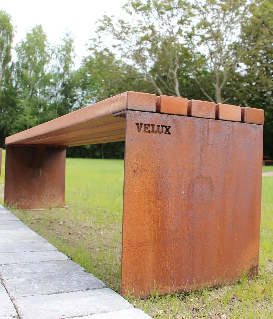 Rustikale Tisch-Bank-Kombination aus Metall mit Holzauflage, in einem grünen Park aufgestellt.