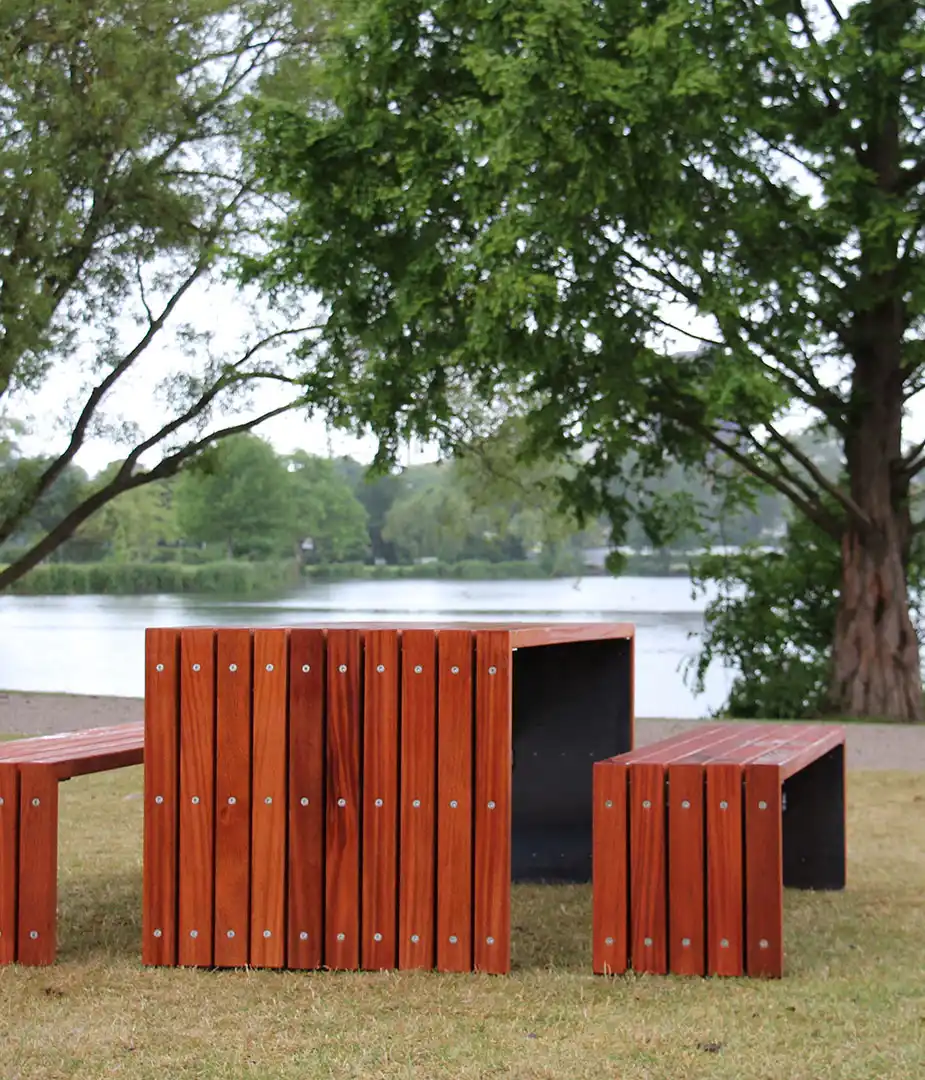 Tisch-Bank-Kombination aus Holz in einer Parklandschaft nahe einem Gewässer, umgeben von Bäumen.