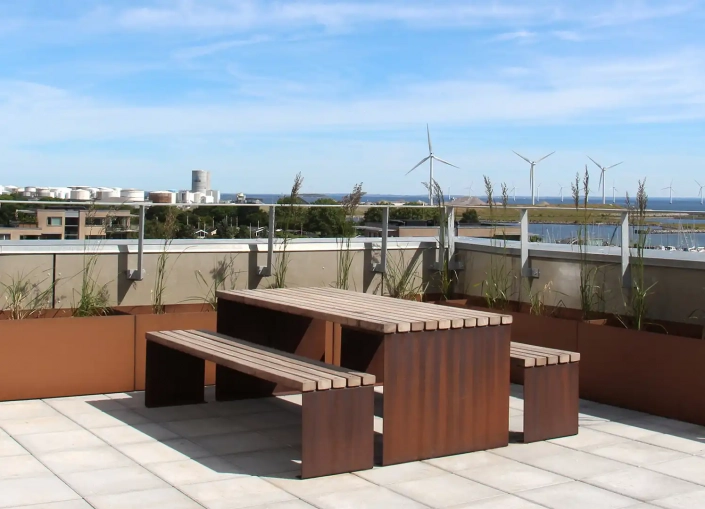 Tisch-Bank-Kombination aus Holz auf einer Dachterrasse, umgeben von Gräsern und mit Blick auf eine Landschaft mit Windkraftanlagen und Wasser.